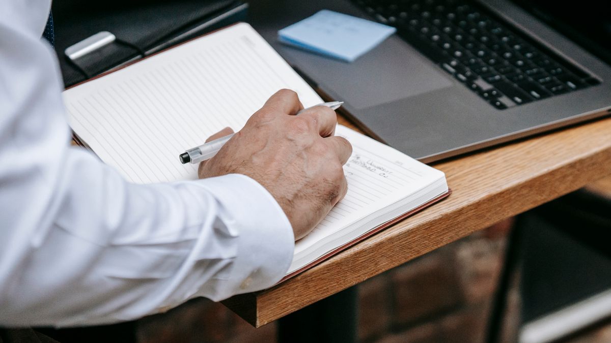 A person in a white shirt writing notes in a lined notebook beside a laptop on a wooden desk.